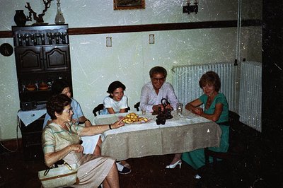 Four people are seated around a table covered with a beige tablecloth. A buffet stands against the wall. The scene suggests a...