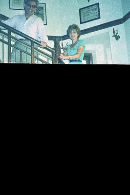 A man and woman pose on an interior staircase. The man, in a white shirt, leans against the railing. The woman wears a turquo...