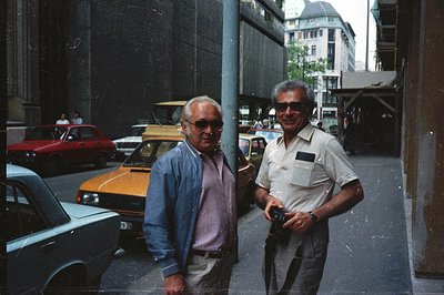 Two men, both with graying hair and sunglasses, pose for a photo on a street lined with older European cars. Appears to be a ...