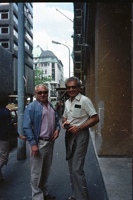 Two men in casual attire, sunglasses, stand in a city street. One wears a denim jacket over a pink shirt, the other a white s...