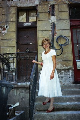A woman in a white midi dress and red heels poses on a weathered stone staircase. Aged building facade behind, showing signs ...