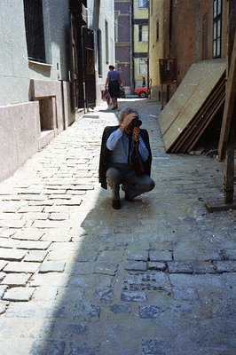 A man in a suit crouches, appearing to photograph the cobblestone street of a narrow, European alleyway. A woman walks away i...