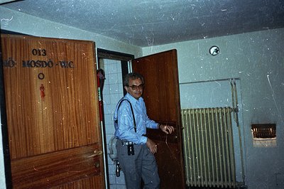 Man exits a doorway marked "Mosdo-WC" (likely restroom signage) on a tiled wall. He wears a light blue button-down shirt, sus...