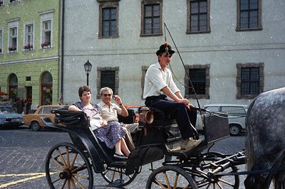 A couple enjoys a horse-drawn carriage ride through a historic European town. Note the traditional driver’s costume and the e...