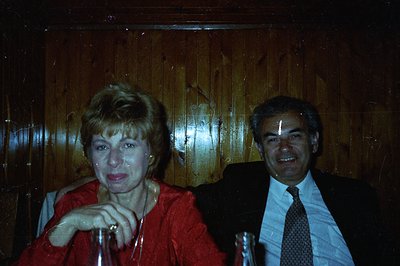 Two individuals, a woman and a man, are captured in a candid portrait against a dark wood-paneled backdrop. The woman wears a...
