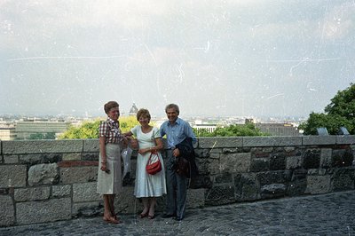 Three figures stand before a stone wall with a cityscape backdrop. The subjects, two women and a man, appear to be on vacatio...
