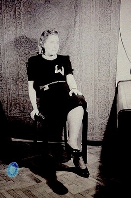 Black and white portrait depicts a woman seated on a wooden chair, dressed in a dark, belted dress and heels. She appears poi...