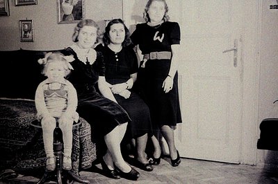A young boy in a stylized costume sits before two women. One wears a dress and lace collar, the other a dark dress with decor...