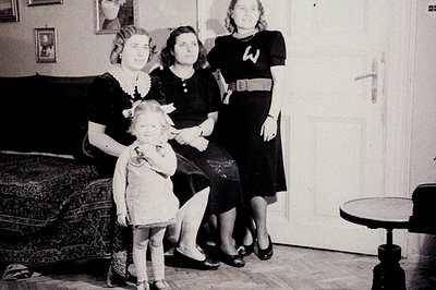 Family portrait, c. 1930s. A young girl stands between two women in formal attire, posed in a parlor. Dark patterned rug, woo...