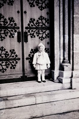A young child stands outdoors before ornate, carved doors. The child wears a heavy, textured winter coat, gloves, and a peake...