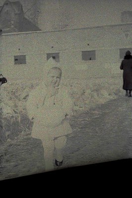Black & white photo showing two figures near a building with small, square windows. One figure, a child in a coat, appears to...