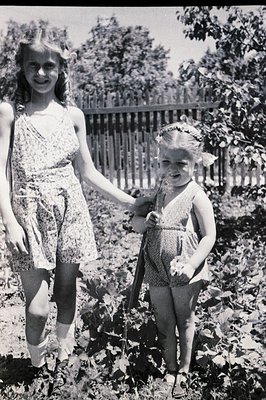 Two young girls, likely sisters, stand hand-in-hand in a garden setting. The elder wears a floral print dress and a flower cr...