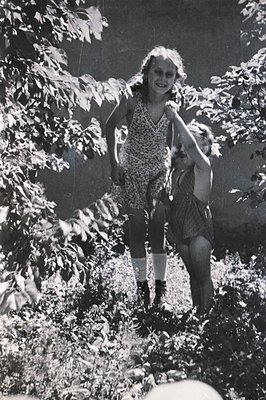 Two young girls playfully navigate overgrown foliage, one appearing to guide the other. They wear vintage summer dresses and ...