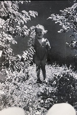 A young child, possibly a girl, stands amidst dense foliage. She wears a patterned, sleeveless dress and a floral headband. T...