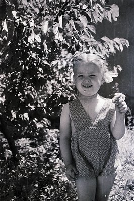 A young girl in a patterned romper smiles directly at the camera, standing in front of a leafy tree. Visible grain and soft f...