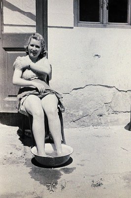 A young woman in a floral dress sits outdoors having her feet soaked in a metal basin. Stone wall and window visible in the b...
