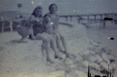 Two figures are faintly visible against a blurred background, likely seated outdoors. The scene exhibits signs of age and dam...