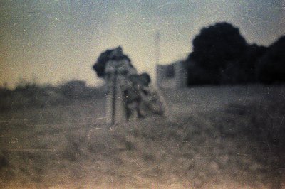 A blurry, monochrome image shows two figures and a tall structure against a dark, indistinct background, likely a forested ar...