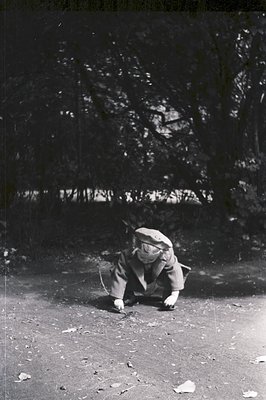 A young child, clad in a short jacket and beret, crawls along a leaf-strewn, gravel path. Background features dense foliage, ...