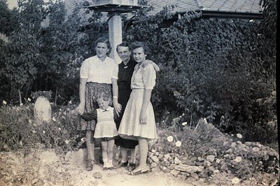 A family portrait: three women and a young girl pose near a stone pillar covered in vines. The women wear 1950s-era dresses a...