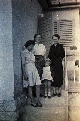Exterior view of three women and a young girl standing on stone steps. Architectural detail: shuttered window, simple façade....
