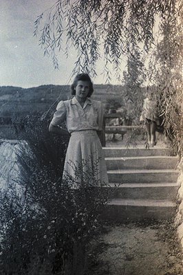 A young woman in a vintage dress stands on stone steps beneath a willow tree. The style suggests the 1950s, evident in the dr...