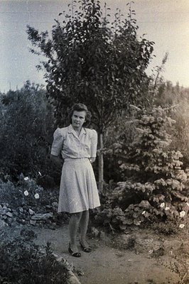 A young woman stands in a lush garden setting, wearing a modest, A-line dress and sandals. The scene features dense foliage a...
