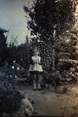A young girl stands in a garden, wearing a simple, knee-length dress and dark shoes. The scene is captured in black and white...