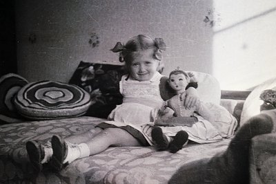 Young girl in pigtails poses on a floral-patterned bed with a doll. A patterned cushion rests behind her. Appears to be a can...
