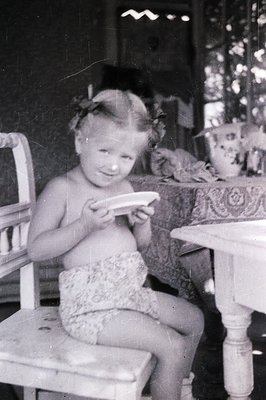 A young girl sits on a chair, holding a small plate; floral patterned fabric shorts are visible. Interior scene, possibly a v...