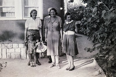A young girl in a patterned dress stands with three women in front of a stone building. The women wear plaid dresses with cin...