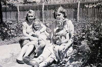 Three young women, possibly sisters, pose outdoors with a rustic wooden fence and lush foliage in the background. The subject...