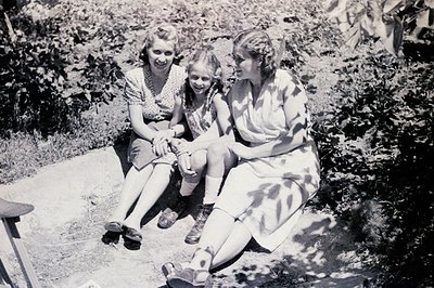 Three women seated in a lush garden setting. The eldest appears to be a mother or guardian, flanking a younger girl and anoth...