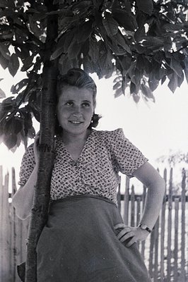 Young woman poses outdoors, leaning against a tree trunk. She wears a printed blouse and full skirt, cinched at the waist. Li...