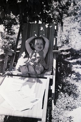 A young child sits in a wooden deck chair, arms raised, with a joyful expression. The chair rests upon newspapers, suggesting...