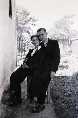 Black and white portrait depicts a formally dressed couple seated on a simple chair beside a wall. Man in pinstripe suit, wom...