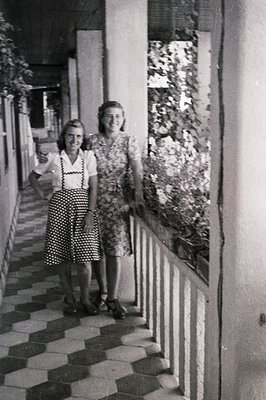Two young women in patterned dresses stand on a patterned tiled terrace with a balcony railing. The architectural style sugge...