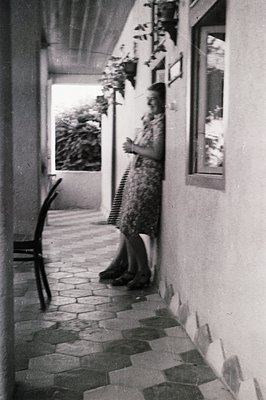 A woman in a patterned dress stands in a tiled courtyard, leaning against a whitewashed wall with potted plants above. An emp...