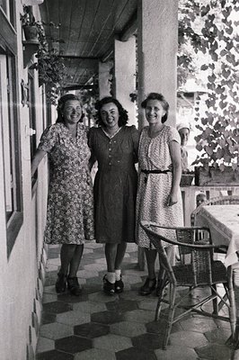 Three women in patterned dresses pose on a tiled terrace. The setting features a stone wall and climbing plants, suggesting a...