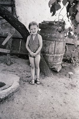 A young girl, possibly 5-7 years old, stands smiling in a rustic courtyard. She wears a patterned romper and sandals. A large...