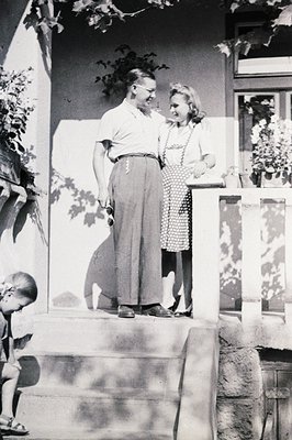 A man and woman stand on a stone staircase, seemingly engaged in conversation. The man wears wide-legged trousers & a tucked-...