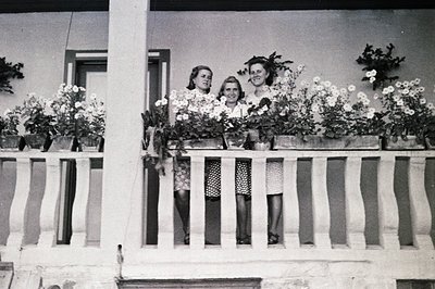 Three women stand on a wrought-iron balcony overflowing with white flowers, likely a coastal resort. Architectural details su...