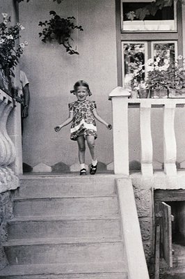 A young girl with a cheerful expression descends a set of stone steps. She wears a patterned dress and Mary Jane shoes. The b...