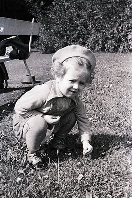 A young child, likely a toddler, kneels in a grassy area, wearing a beret and corduroy clothing. The child appears to be reac...