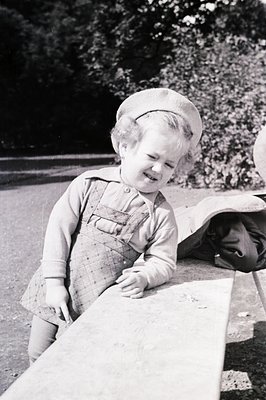 A young child in overalls and a patterned hat leans against a stone bench, seemingly amused. Likely a candid portrait, the se...