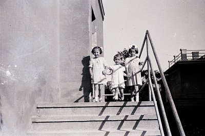Three young girls in matching dresses and bows pose on wide concrete stairs, shadows extending from the building's façade. A ...