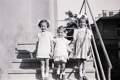 Three young girls, each wearing a smocked dress and Mary Jane shoes, stand on concrete steps. The girls have short, curled ha...