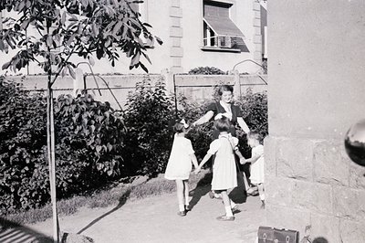 Four children, three in matching dresses, and a woman appear to be playing a game, holding hands and moving forward. A partia...