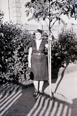 A young woman stands holding a newly planted tree sapling, her gaze directed forward. She wears a belted, knee-length dress, ...