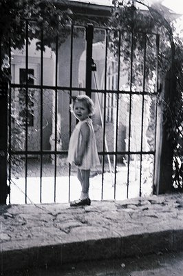 A young girl stands framed by a wrought-iron gate, likely in a garden or courtyard setting. She wears a simple, short-sleeved...
